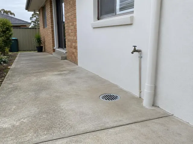 Outdoor concrete paving beside a brick and rendered house wall with a round drain grate in Lalor Park