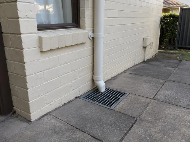 Stormwater grate beside a cream brick house wall and downpipe on dry paving in Blacktown